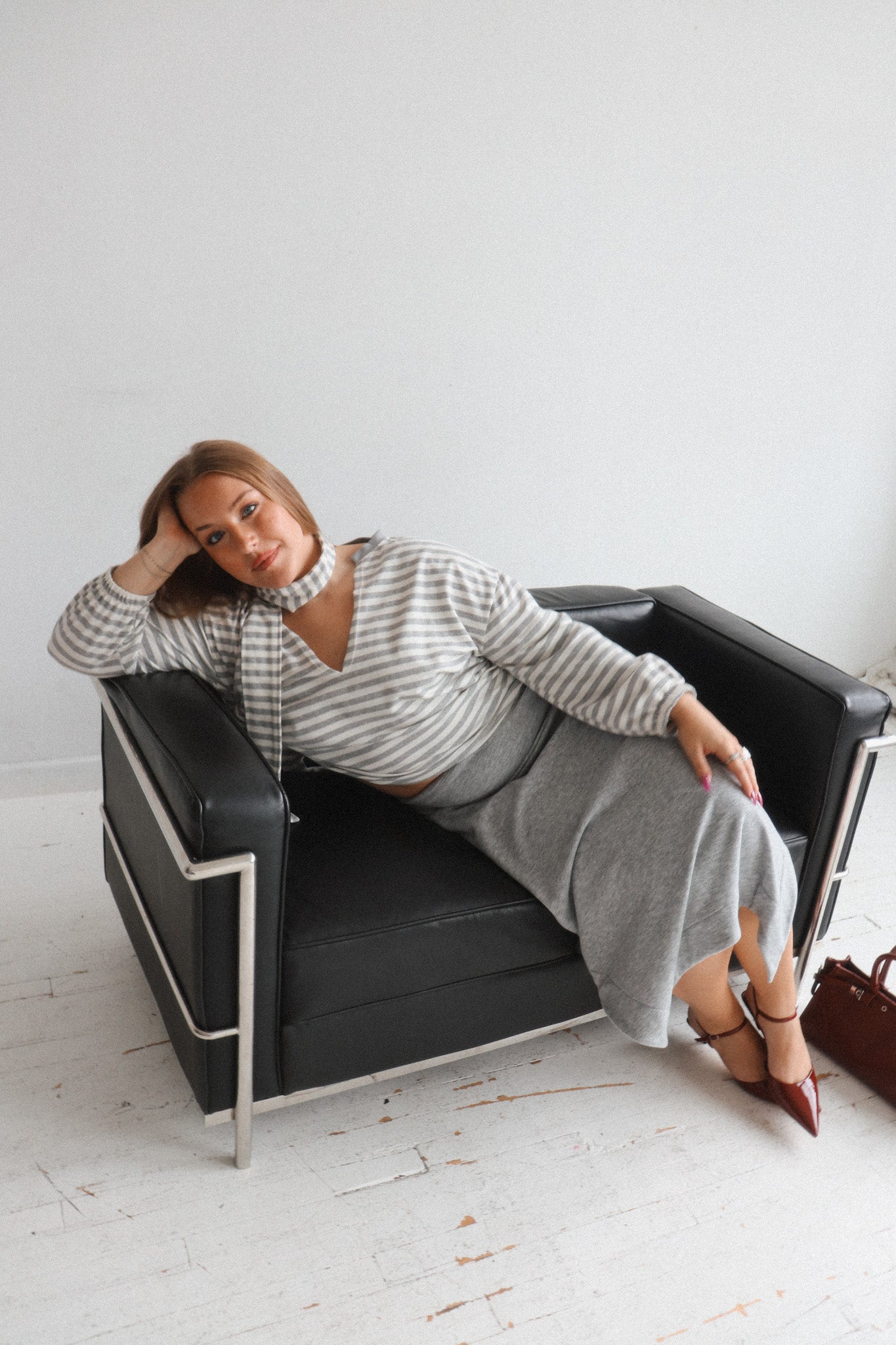 A woman reclining on a black leather armchair with a white wall and white floor in the background, wearing a gray and white striped long-sleeve top and a gray midi skirt.