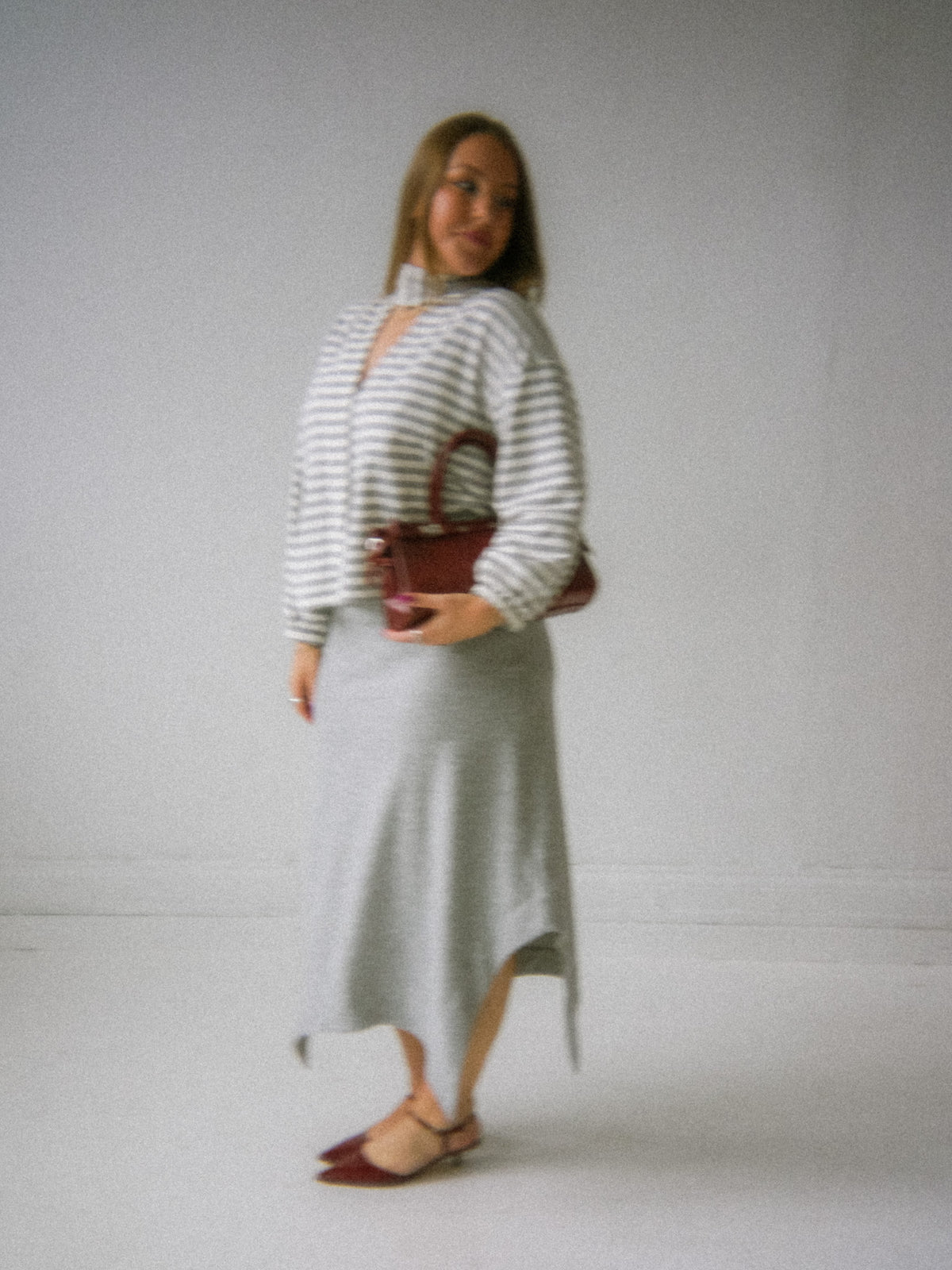 A woman modeling a gray and white striped mock-neck long-sleeve top with a keyhole cutout, paired with a light gray midi skirt and holding a brown handbag.