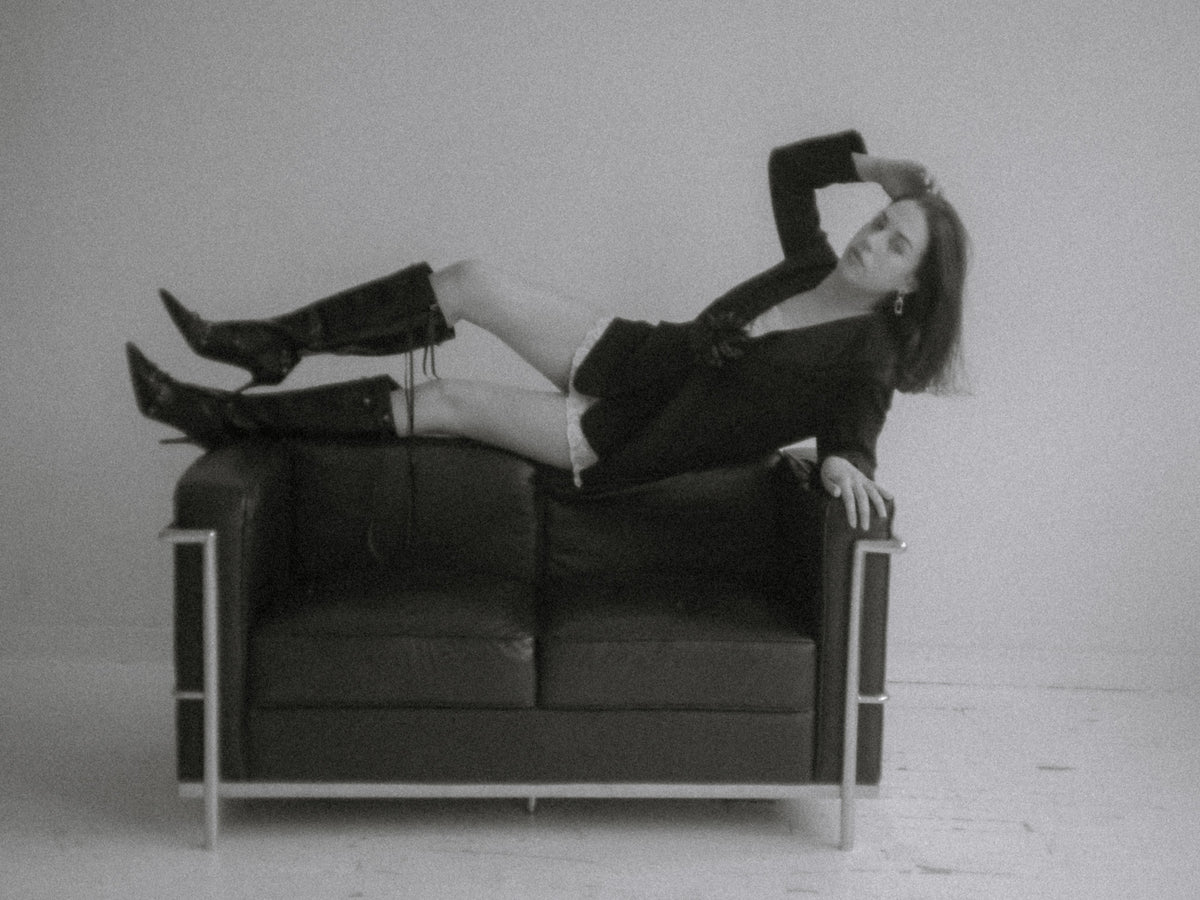 A black-and-white fashion photo showing a woman reclining across a dark leather sofa in a minimalist studio setting.
